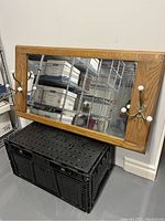 Oak framed rectangular mirror leaning against a white wall, resting on black storage bins. Two brass hooks with white porcelain knobs are mounted on one side of the frame.