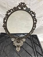 Front view of an antique round mirror with an intricately detailed metal frame and stand placed on a black plastic grid surface against a white backdrop.