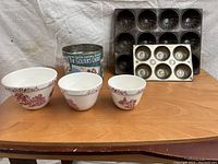 Photo showing set of 3 ceramic mixing bowls with red decorative pattern, 2 metal muffin tins with six muffin cups each, and a vintage cylindrical tin container.