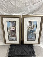Pair of tall rectangular framed pictures depicting storefronts, one with a red and cream awning and one with a blue and cream awning, photographed against a white backdrop.