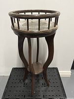 Wooden planter stand with circular railing top and four curved legs with a small base shelf.