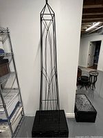 Photo of a free standing black metal plant trellis against a white wall, showing full height and detailed geometric design