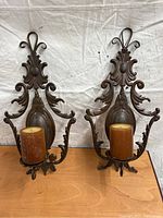 Front view of two ornate metal wall hanging candle holders each holding a brown pillar candle, set against a fabric backdrop