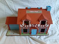Top front angle view of vintage Fisher Price Little People Neighborhood playhouse showing red roof with dormers, blue front door, window openings, and attached garage.