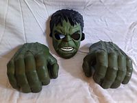 Front view of green Hulk mask and both green Hulk fist gloves laid out on white fabric background.