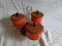 Three orange ceramic condiment holders with teak lids mounted in a teak and metal caddy, viewed overhead on a fabric surface.