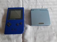 Original front view of blue Gameboy Pocket and closed silver-blue Gameboy Advance SP on fabric background