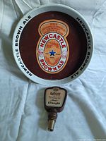 Photo of Newcastle Brown Ale metal tray with Labatt's beer tap handle in front on white fabric background.