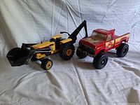 Photo showing both the yellow backhoe loader and red pickup truck side by side on white fabric background, highlighting their size and condition