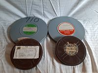 Four vintage film reels and canisters laid out on fabric background, viewed from above, showing labels and different sizes.