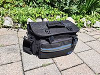Closed black camera carrying bag with blue stripe and handle, placed on stone patio outdoors.