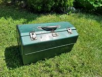 Closed vintage green metal fishing tackle box on grass showing black handle and metal latches