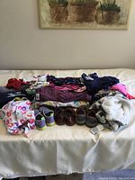Wide view of the assortment of toddler clothes and shoes spread out on a bed, showing variety of items and three pairs of shoes included.