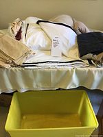 Bedding items arranged on a bed including folded beige sheets and pillowcases, white sheets, duvet cover with black trim, and a dark gray folded blanket visible on right side.