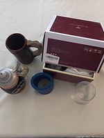 Photo showing brown crockery beer stein with wooden handle, ceramic beer stein in original box, blue foam bottle cozy, and one empty clear glass tumbler.