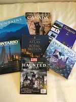 Seven hardcover coffee table books displayed on a bed, titles visible including Mountains, The Wildlife of Canada, Ontario, Atlas Royal Britain, Oak Ridges Moraine, and Haunted Places.