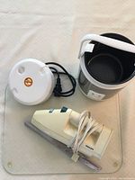 Mini rice cooker with removable inner pot and white handle, electric carving knife with cord, and glass cutting board all visible from above on white surface.