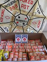 Photo of Rummoli game board backdrop with box of stacked red and white poker chips and two Bicycle decks visible in front.