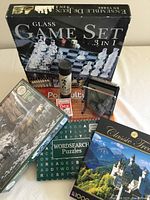 Photo showing the glass 3-in-1 game set box, cards, pick up sticks, word search book, Trivial Pursuit DVD, and pin art toy arranged on white surface.