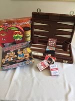 Leather cased backgammon set displayed open with dice, chips, and instructions; boxed card and board games including Trivial Pursuit DVD pop culture edition, Apples to Apples family edition, and Hedbanz game; Uno card deck and two poker playing card decks.