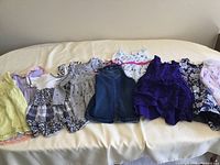 Set of nine girl's dresses in various colors and styles arranged on a bed; showing variety including sleeveless, long sleeved, lace, denim, floral patterns