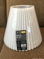 White pleated lamp shade in original plastic with label, displayed on brown fabric background
