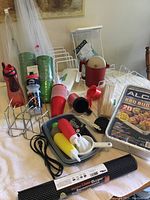 Photo showing various kitchen and BBQ related items including packages of disposable BBQ trays, water bottles, red and green plastic cups, metal serviette holders, skewers, plastic kitchen utensils, and grip shelf liner.