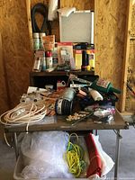 Full lot showing assorted household and hardware items arranged on and under a folding table inside unfinished wooden room.