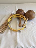 Photo showing five wooden drumsticks, one yellow plastic tambourine with metal jingles, and two wooden maracas on a white surface.