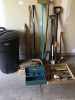 Overall view of tools and storage containers including garbage pail, tackle box, garden tools, mop, and hand tools in boxes on floor