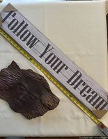 Photo showing 'Follow Your Dream' wooden wall plaque alongside the woman face sculpture with measuring tape for scale.