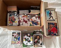 Photo showing two boxes filled with hockey trading cards and multiple stacks of loose hockey cards on a white cloth surface.