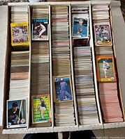 Top view of a large box divided into 5 sections, each filled with stacks of baseball cards. Several cards are placed on top showing various players and card brands.
