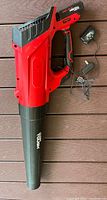 Red and black cordless leaf blower laying on wooden deck. Includes attached rechargeable battery and separate charger with cables.