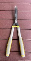 Metal garden shears with black blades and extendable gray and yellow handles laid flat on a red wooden deck.