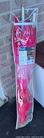 Garden umbrella packed inside a clear zipper storage bag leaning against a brick wall, showing red fabric and wooden handle visible at the top.