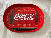 Front view of an oval vintage Coca-Cola serving tray with red background and two classic Coke bottles, along with Coca-Cola branding and decorative gold designs.