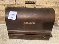Closed view of the dark wooden box with hinged lid featuring 'Singer' gold lettering on front. Shows surface wear and scratch marks.