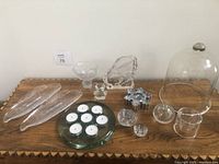 Collection of assorted glass items displayed on a wooden surface including clear glass fish-shaped dishes, flower shaped bowl, candle holder with tea lights, small glass rings, glass dome, and others.