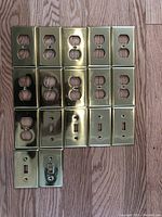 Top view of brass plated outlet and light switch covers arranged on wood floor, total of 17 covers.
