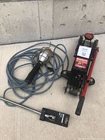 Motomaster 2 ton hydraulic trolley jack with red handle lever, black body, wheels, accompanied by handheld trouble light with long blue cord and protective cage around the bulb.