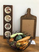 Photo of carved faux gourds displayed in the metal twig basket, with wooden push-pin board and a panel with four set embroidered art pieces in background
