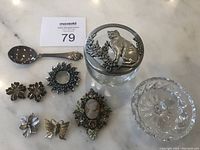 Photo of all items together: Gerber baby spoon, vintage brooches and pins including a cameo brooch, floral and butterfly designs, a round glass container with a pewter lid featuring a cat and floral motif.
