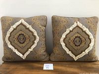 Pair of two square pillows with central diamond-shaped layered fabric design in beige and gray colors, featuring fringe and bead embellishments, placed side by side against a white background.