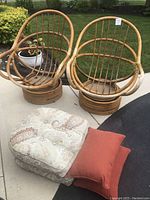 Two rattan chairs with circular backs and bases, one large patterned cushion and one smaller orange cushion on the ground in front