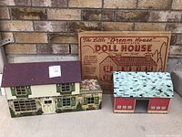 Photo showing vintage steel doll house, steel barn, and boxed Eatons cardboard doll house together against a brick wall