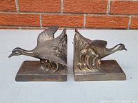 Pair of antique silver plate Art Deco bookends featuring raised relief flying geese designs on rectangular bases against brick background.
