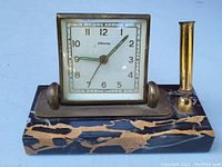 Front view of antique Blessing desk clock with brass frame, showing white dial with gold Arabic numerals and hands, mounted on black and brown marble base with brass pen holder attached right side