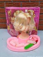 Front angled view of Barbie Beauty Center styling head with hair rollers, combs, pink base, and original box in background.