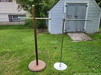 Photo of two clothing racks, one wooden with round base and brass ends, and one chrome metal with round base, standing outdoors on grass near a shed.